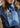 At Home With Ray Cactus Rose Silky Ribbon Scarf in July Sky shown close up at the neck on a woman in a denim western shirt, the watercolor-washed blue tones and antique gold illustrated motifs adding an ethereal southwestern elegance to the look.