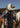 At Home With Ray Cactus Rose Silky Ribbon Scarf in Blue Corn deep indigo navy shown close up at the neck on a woman in a western vest and white hat, with a horse and lasso visible in the background.